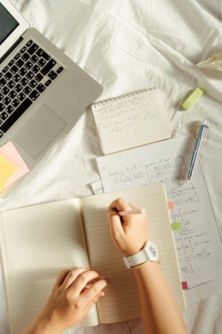 Hands writing math formulas in a notebook with a laptop and stationery on the bed.